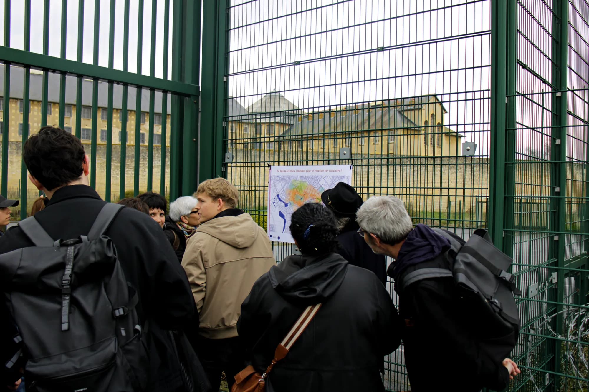 People lean towards the map on the fence, in front of the prison buildings