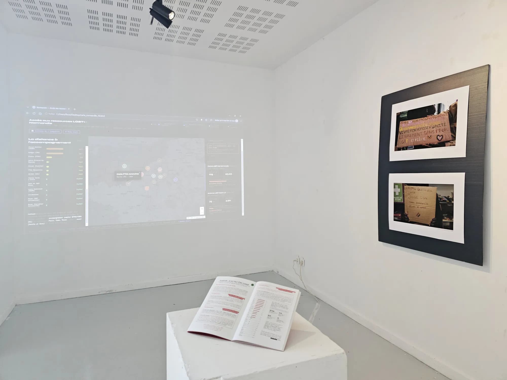 Installation view: an interactive map projected on the wall, a panel of photographs and a booklet open on a plinth