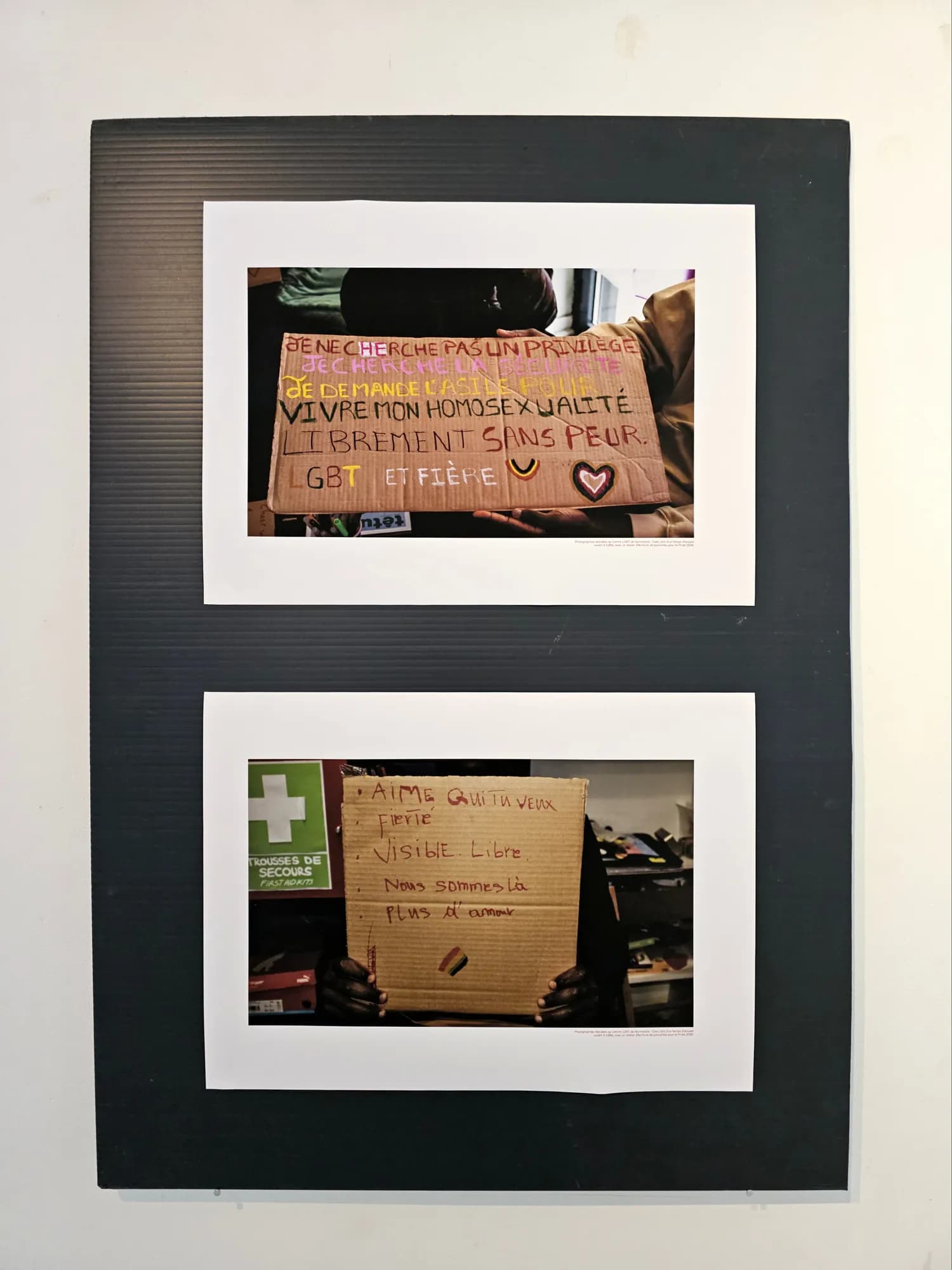 Black panel with two framed photographs of handwritten cardboard placards, one reading “Je ne cherche pas un privilège, je veux vivre mon homosexualité librement sans peur”