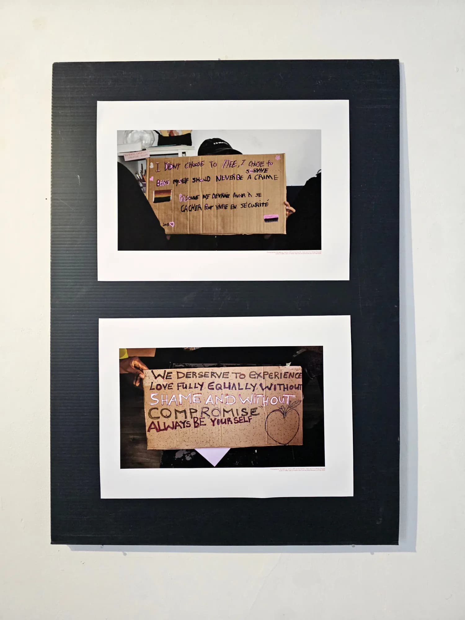 Black panel with two framed photographs of handwritten cardboard placards, one reading “We deserve to experience love fully, equally, without shame or compromise”